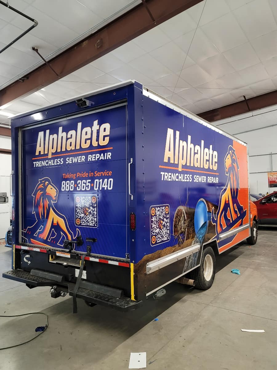 Custom vinyl wrap on a box truck featuring "Alphalete Trenchless Sewer Repair" branding, showcasing a lion logo, contact information, and a vibrant blue and orange color scheme, emphasizing mobile advertising for businesses.