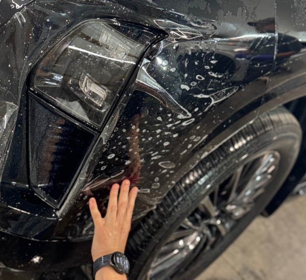 Hand applying paint protection film on a black vehicle's fender, showcasing bubbles and water, emphasizing vehicle care and protection against environmental damage.