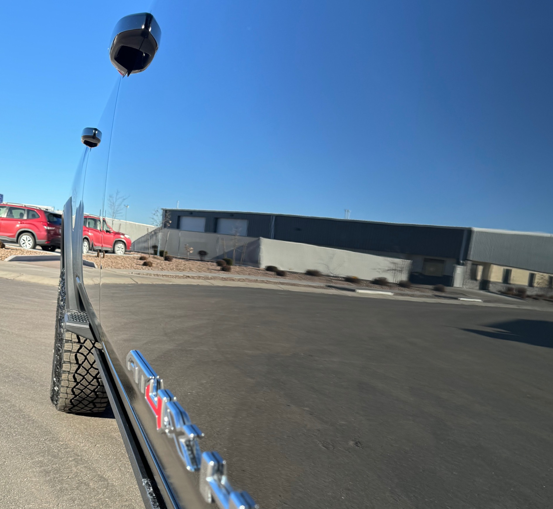 Reflective side view of a vehicle showcasing a glossy finish, highlighting the quality of auto detailing services offered by Luxe Auto Salon, with a blue sky and a red vehicle in the background.